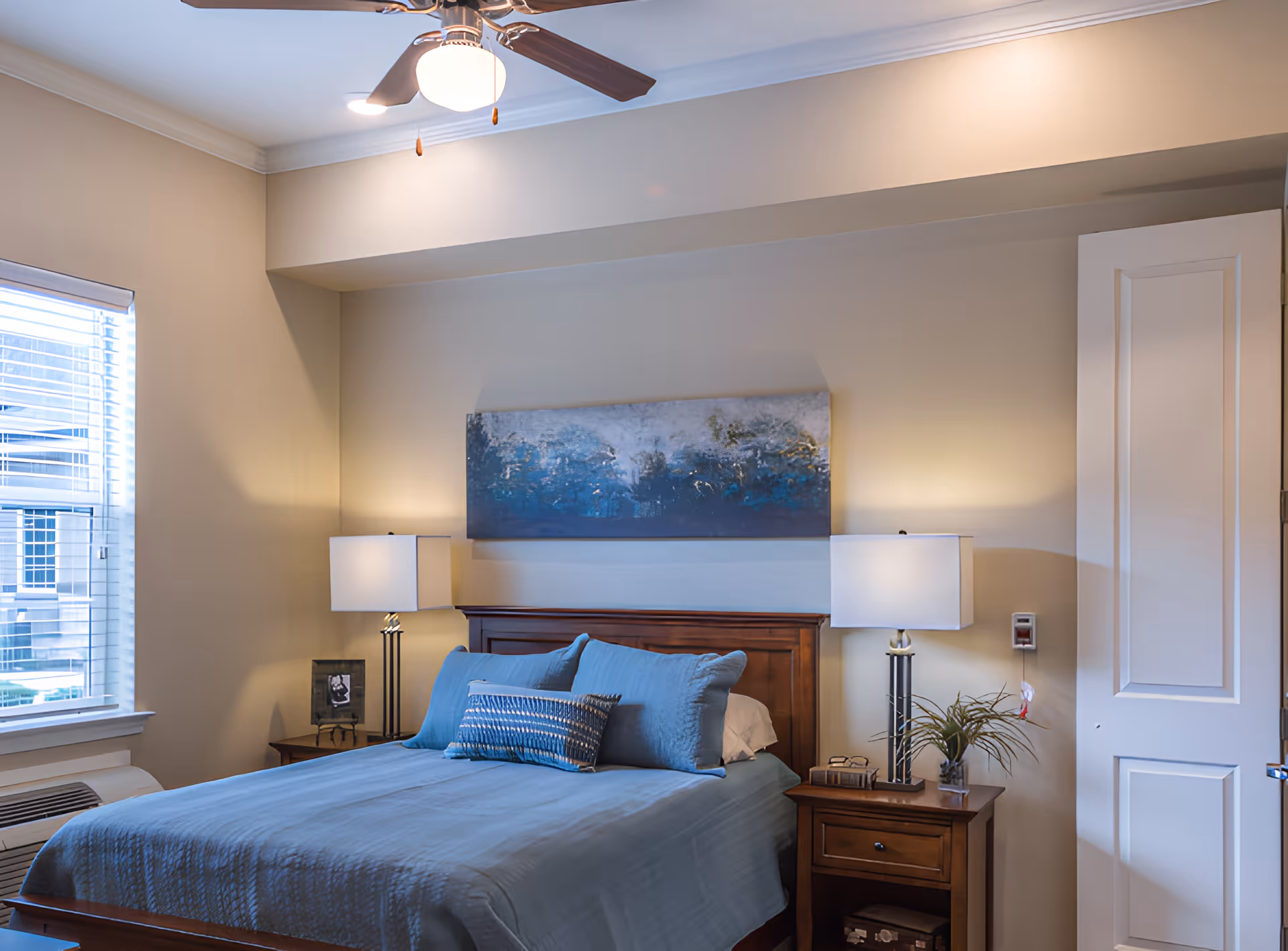 A bedroom with a wooden bed frame and blue bedding, two matching bedside tables each with a lamp, a window with blinds on the left, a ceiling fan with light, and a large abstract painting above the bed.