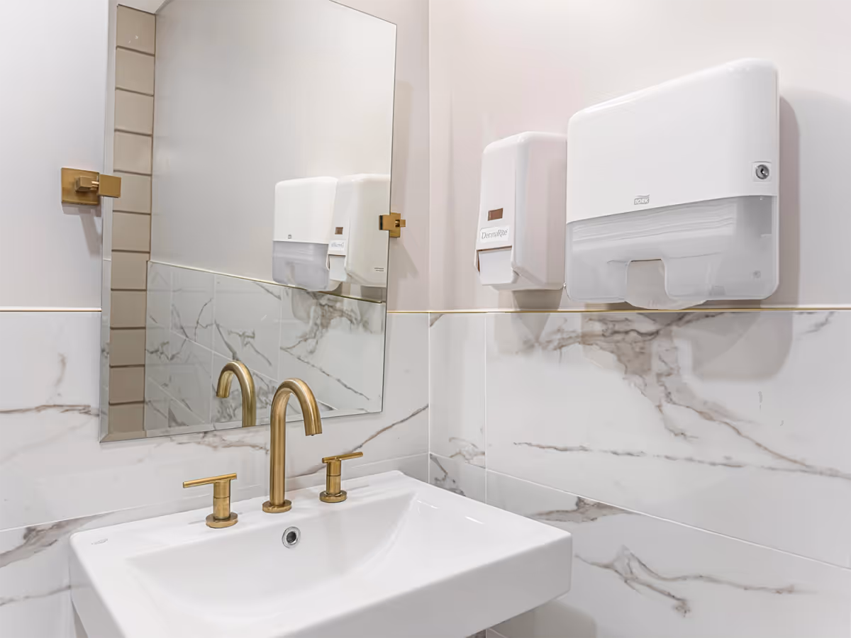 A clean bathroom with a marble-tiled wall, a white sink, and gold-finished faucets with wall-mounted soap dispensers and a mirror.