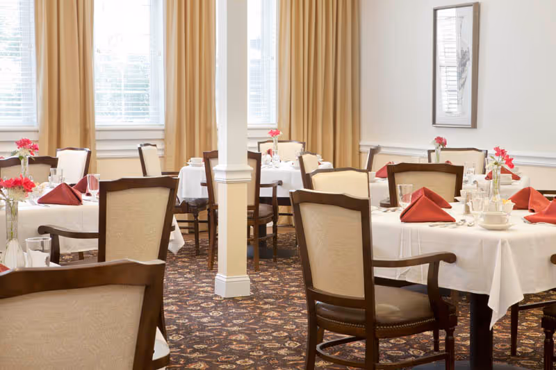A bright dining room with multiple round tables covered with white tablecloths, each set with red folded napkins, glassware, and small vases holding pink flowers. The room has beige curtains, patterned carpet, and framed artwork on the walls.