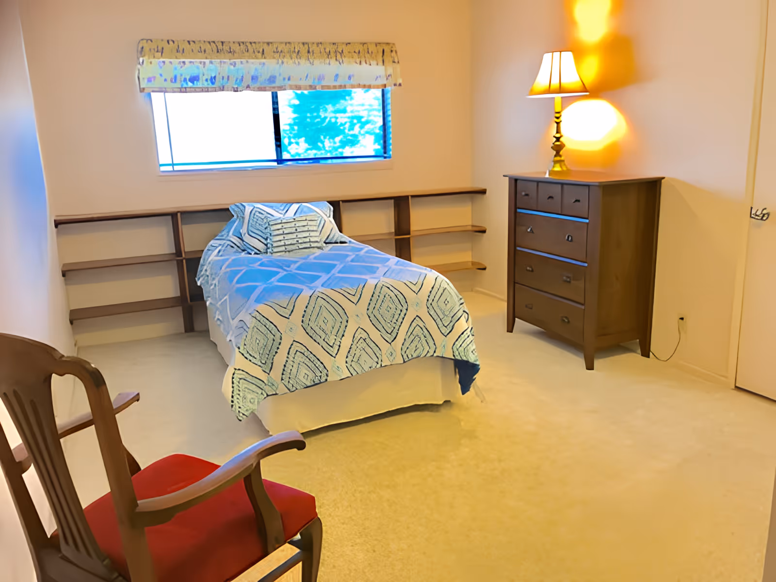 A simple bedroom with a single bed covered in a patterned quilt, a wooden dresser with a lamp, and a wooden chair.