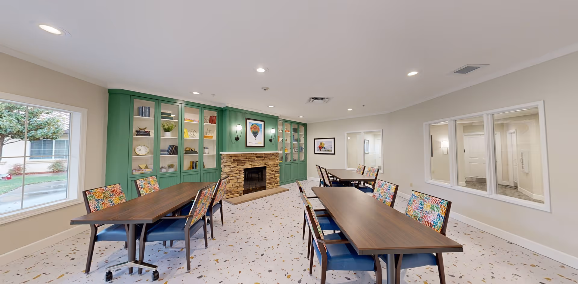 A bright and spacious common room with two wooden tables and colorful cushioned chairs. The room features a stone fireplace with a green built-in bookshelf on either side, decorated with books and plants. Large windows allow natural light to enter, and the floor has a terrazzo pattern. The walls are light-colored, and there are framed pictures hanging above the fireplace and on the adjacent wall.