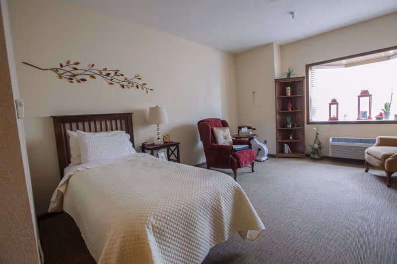 Sunny assisted-living bedroom with a single bed, nightstand, a red armchair, bookshelf and a large window.