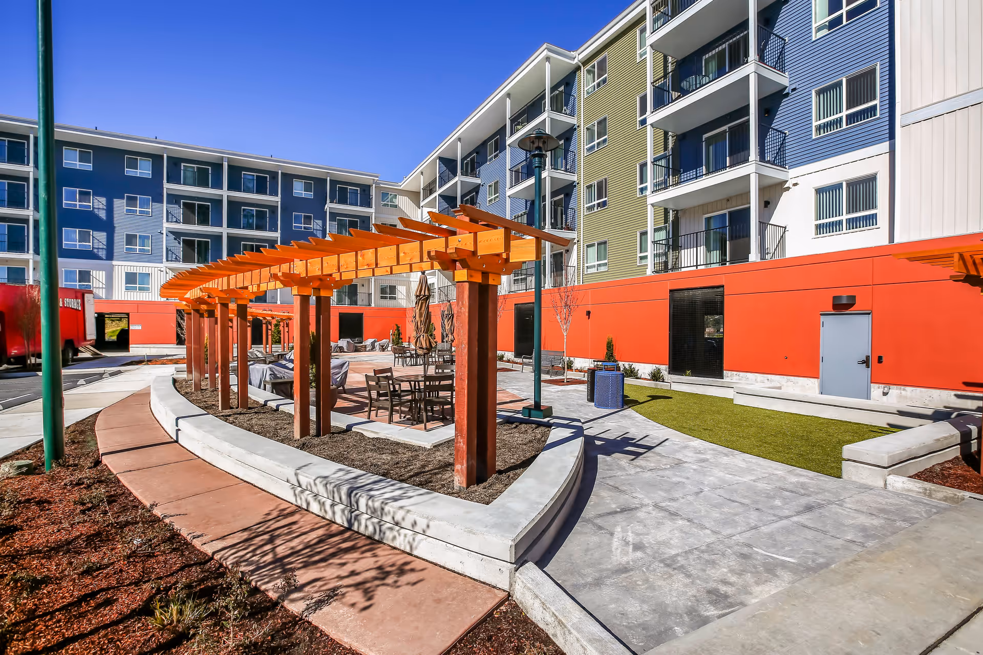 Outdoor courtyard area of a senior living facility with a wooden pergola, patio tables and chairs, concrete walkways, and a multi-story building with balconies in the background under a clear blue sky.