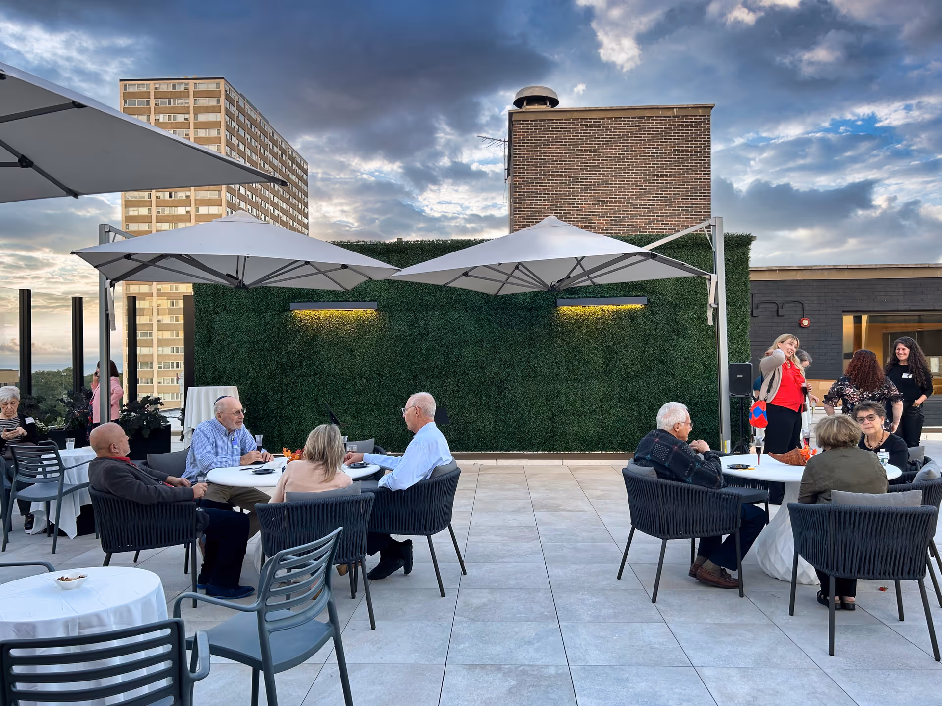 Outdoor patio area at The Selfhelp Home Senior Living Community with several elderly people sitting at round tables under large white umbrellas, engaging in conversation. The patio has tiled flooring, a green wall with lighting, and a tall brick structure in the background. The sky is partly cloudy during sunset.