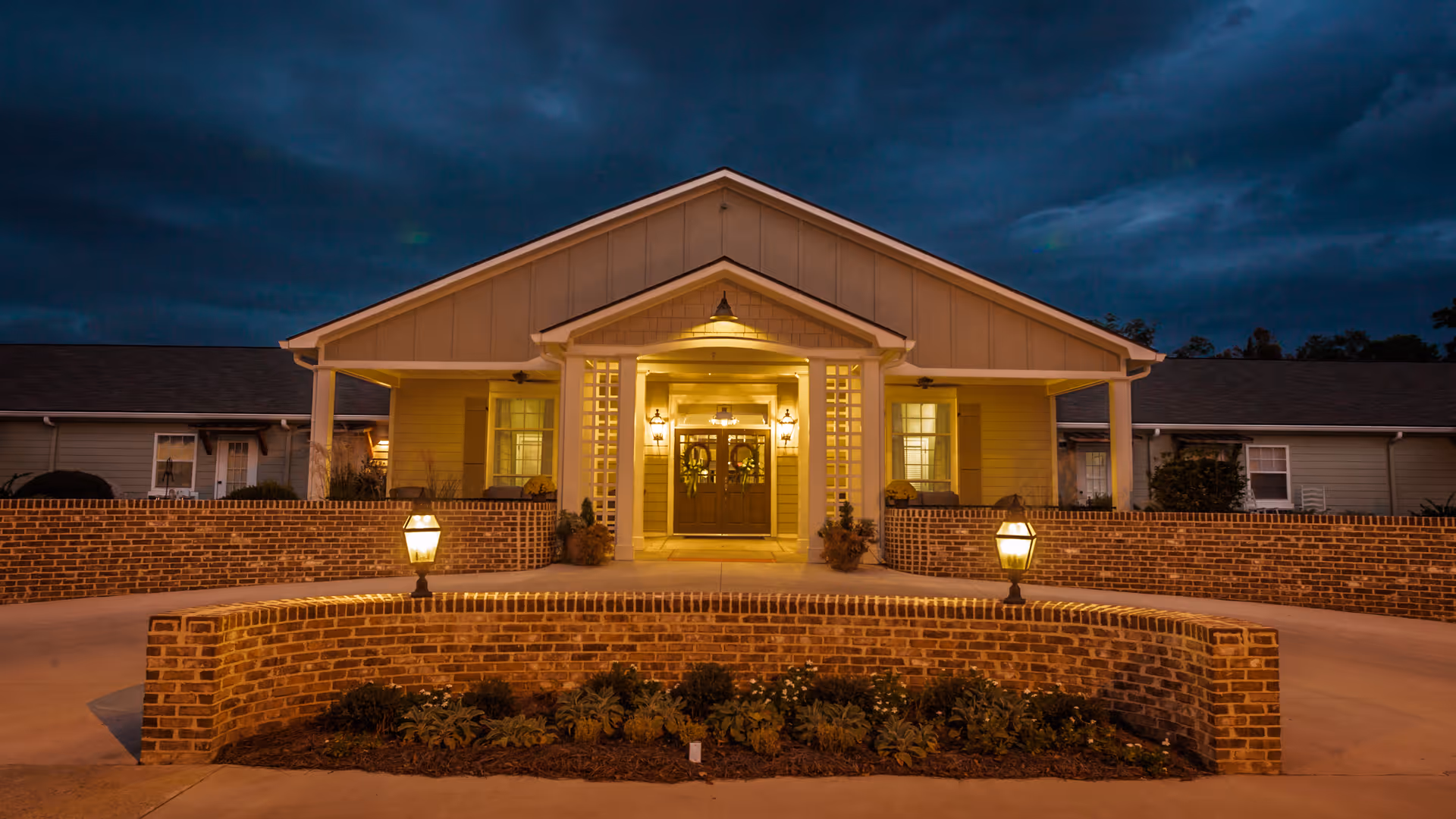 Front exterior view of Fellowship Cordelia Manor Assisted Living Community at dusk, showing a well-lit entrance with double doors, brick walls, and two lantern-style lights on either side of a curved brick planter with greenery.