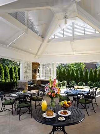 Covered outdoor dining patio with round tables set for meals, a floral centerpiece, and surrounding greenery.