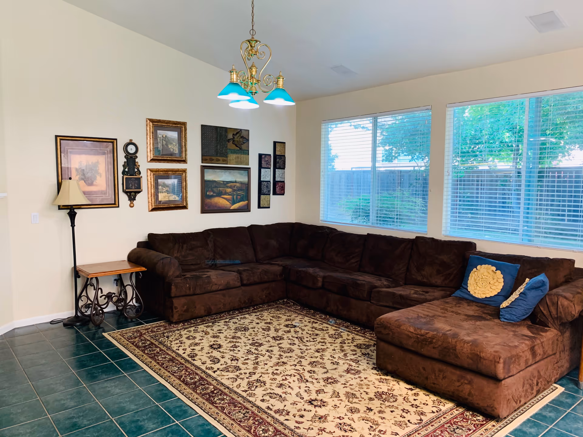 A cozy living room with a large brown sectional sofa adorned with two decorative pillows. The room features a patterned area rug on a green tiled floor, a side table with a lamp, and several framed artworks and a clock on the wall. Large windows with white blinds allow natural light to brighten the space.