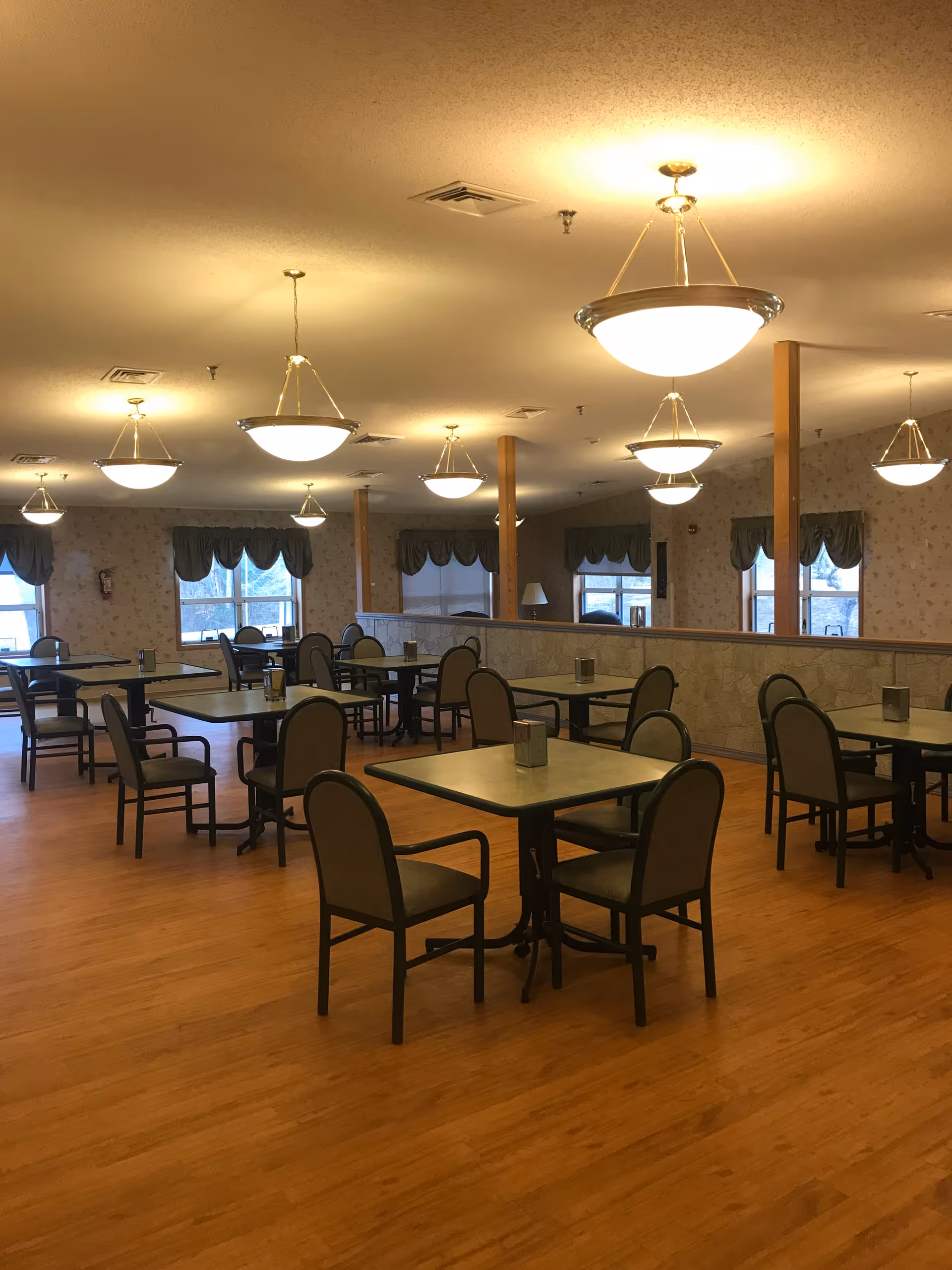 Spacious dining room with multiple square tables and chairs on a wood floor under hanging ceiling lights.