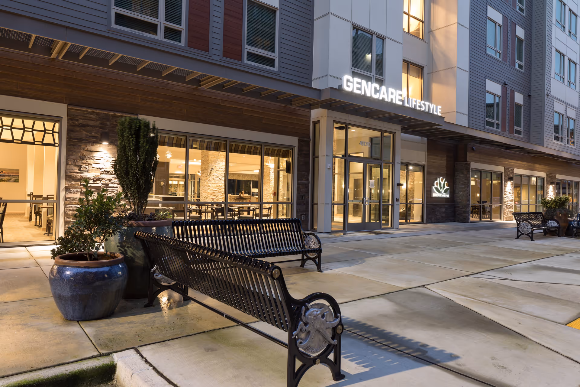 Evening exterior view of GenCare Lifestyle Tacoma at Point Ruston showing the building entrance with large glass doors and windows. There are black metal benches and large potted plants on the sidewalk in front of the building. The interior lighting reveals a dining area inside.