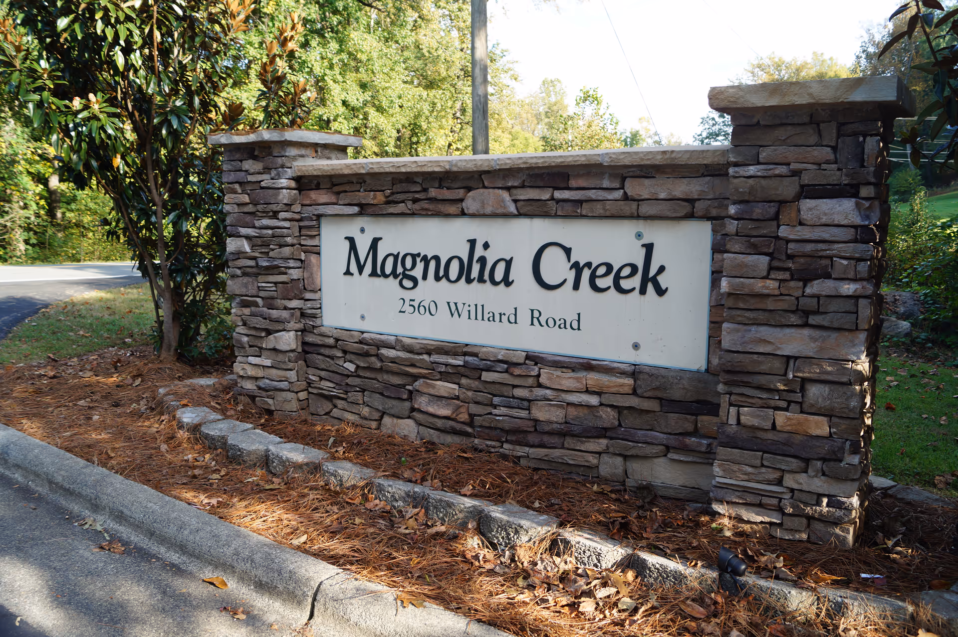 Stone entrance sign for "Magnolia Creek" with the address 2560 Willard Road beside a road and trees.