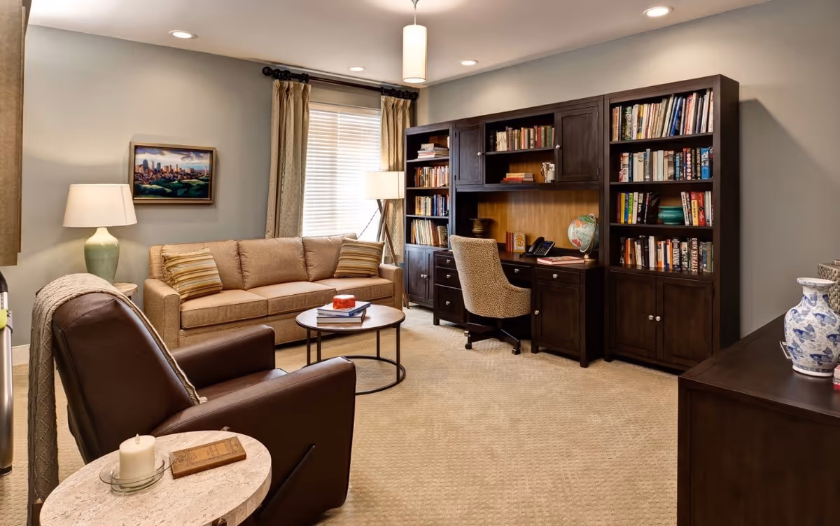 A cozy living room with a beige sofa, a brown leather armchair, a round coffee table with books, a large dark wood bookshelf with books and a globe, a desk with a chair, two lamps, and a window with beige curtains.