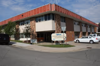 Exterior view of a two-story brick and concrete building with a red roof trim, identified as Uinta Senior Citizens Evanston Adult Day Care by a sign in front. Several cars are parked near the building on a paved street under a partly cloudy sky.