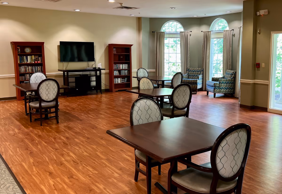 Spacious common room with wood floors, several tables and upholstered chairs, a TV with bookshelves, and seating by tall arched windows.