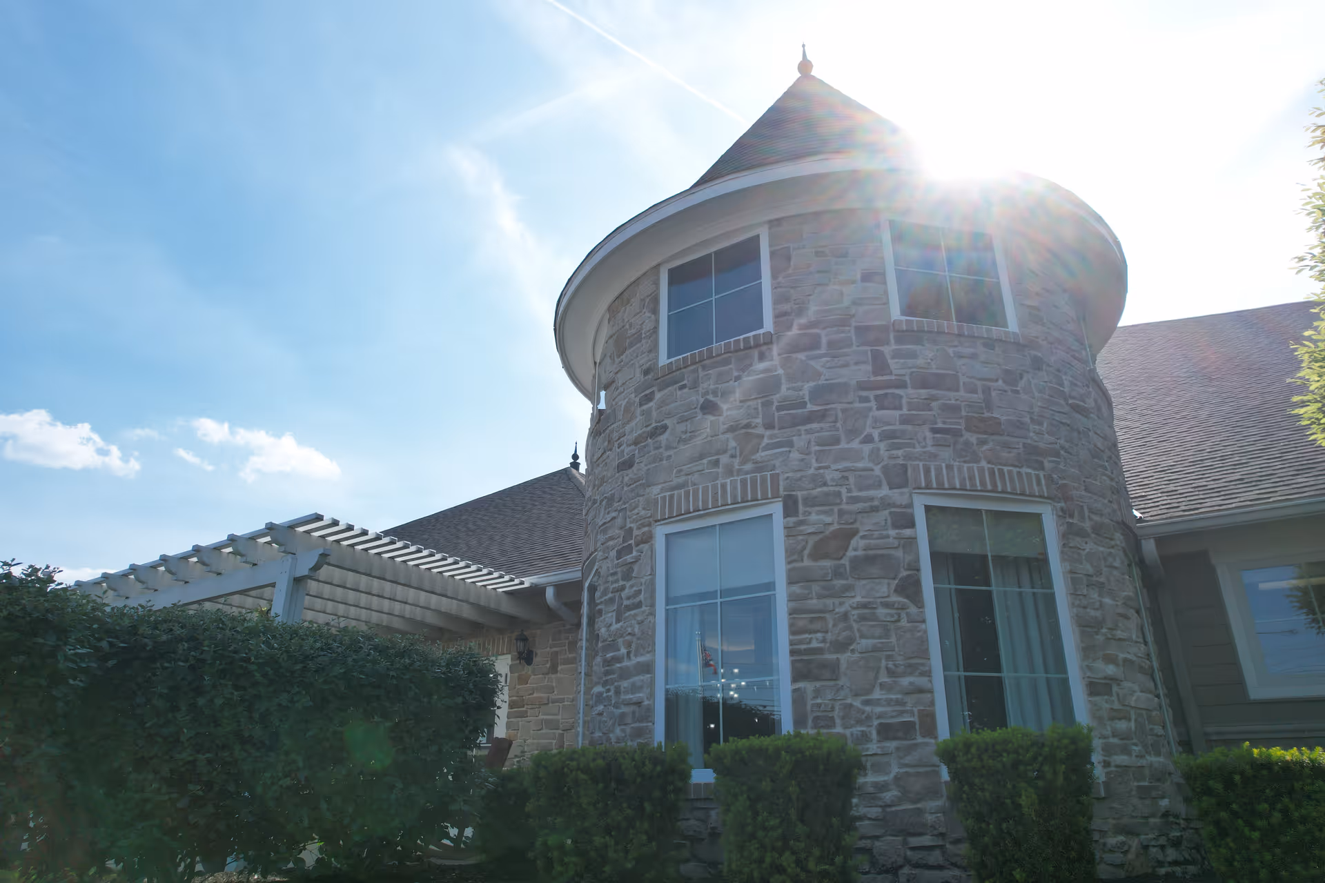 Exterior view of a senior living facility building with a stone facade and a turret-like rounded section with windows. There is a pergola on the left side and neatly trimmed bushes in front of the building. The sun is shining brightly in the sky above.