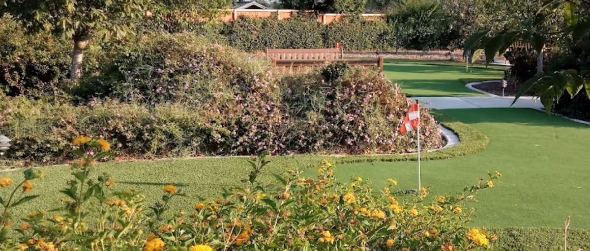 A well-maintained outdoor garden area with a putting green featuring a small red and white checkered flag. There are various green bushes, flowering plants with yellow and purple flowers, and a wooden bench in the background. Trees and a fence are visible further behind the garden.