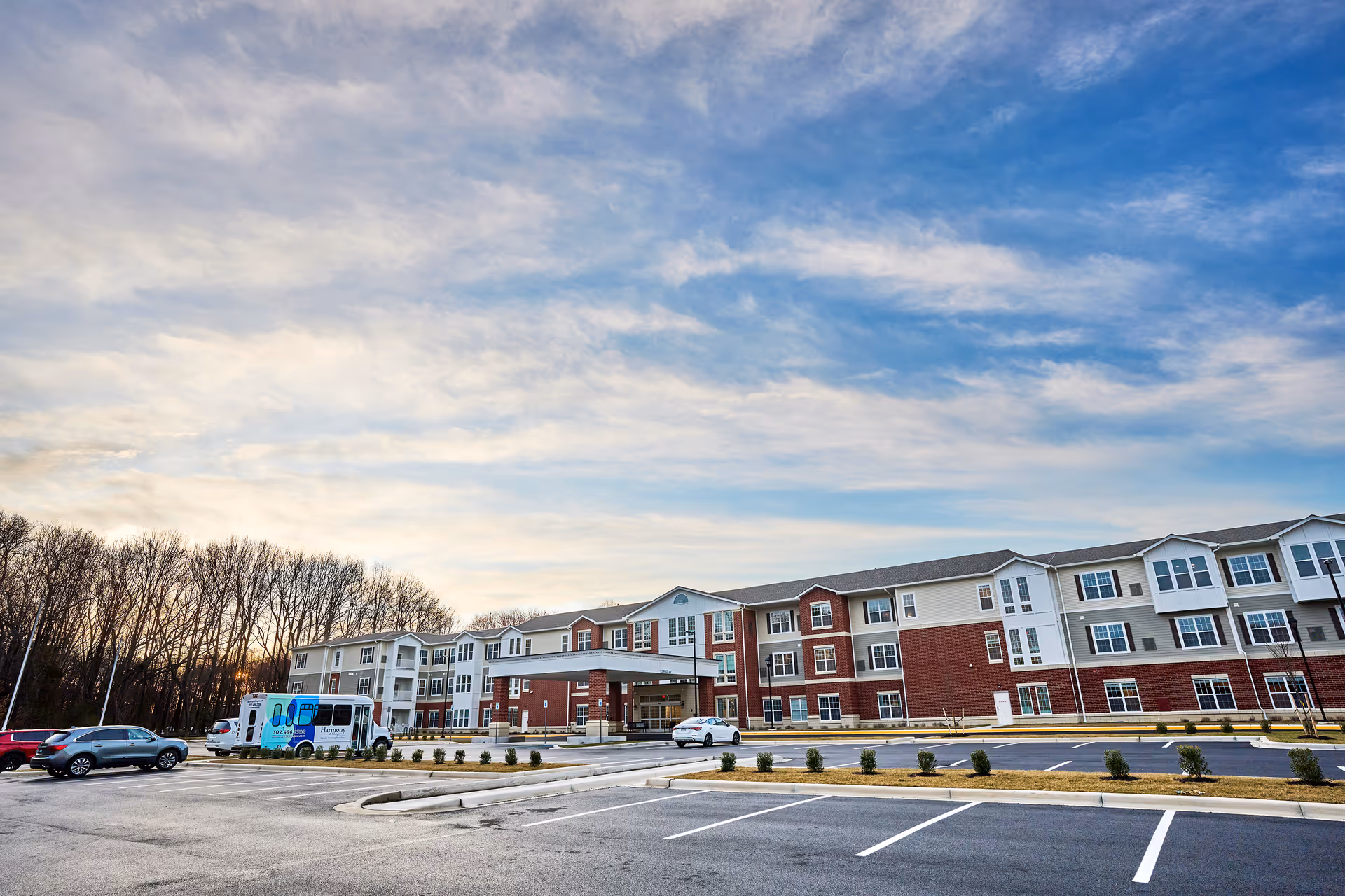 Front exterior of a three-story senior living building with a parking lot and shuttle bus under a cloudy sky.
