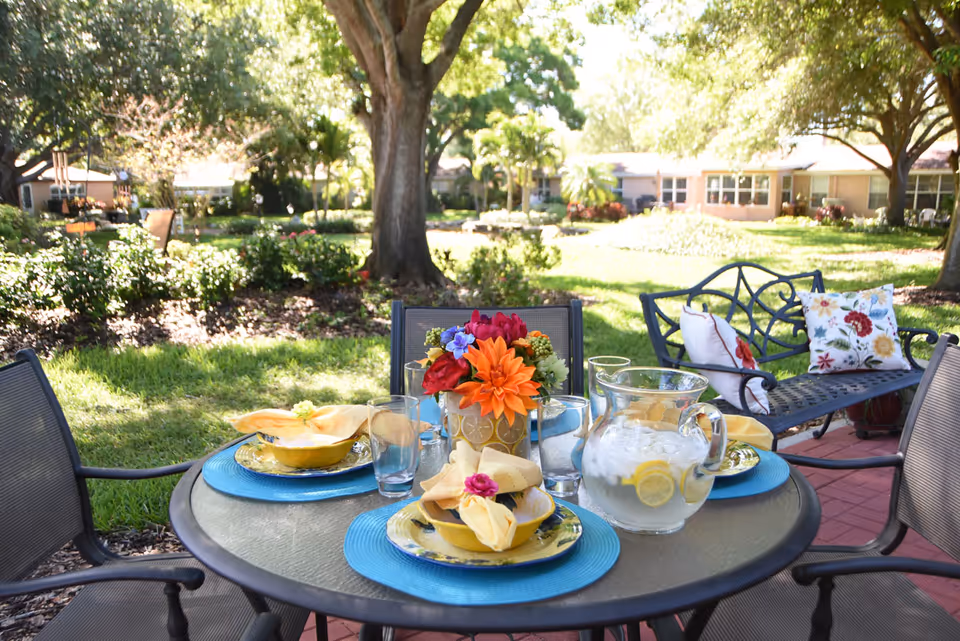 Outdoor patio dining table set with colorful place settings, a pitcher of lemonade and a floral centerpiece on a lawn with trees and buildings in the background.