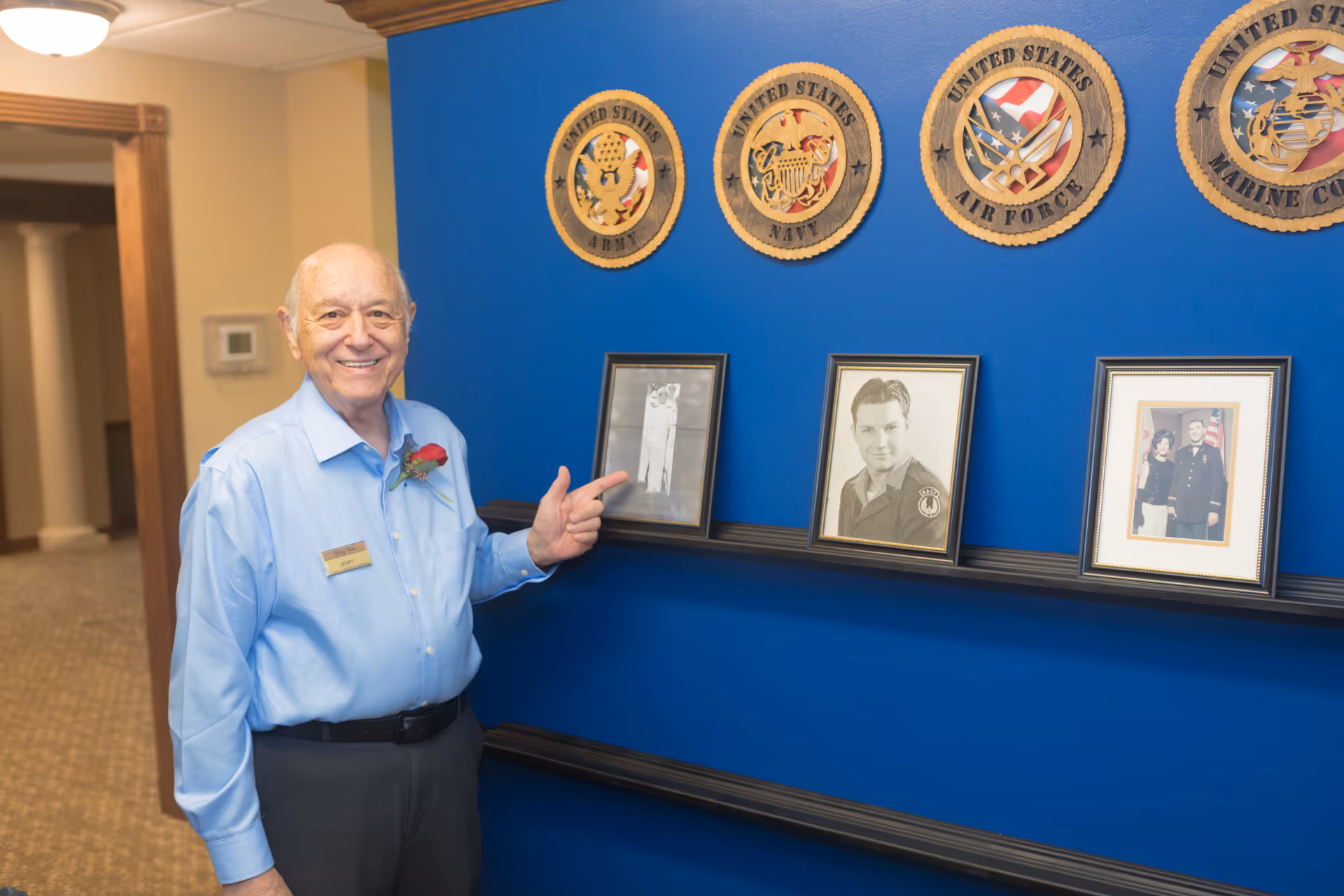 An elderly man wearing a light blue shirt with a name tag and a red rose pinned to his shirt stands smiling and pointing at framed photographs on a blue wall. Above the photos are wooden plaques representing the United States Army, Navy, Air Force, and Marine Corps. The setting appears to be an interior hallway of a retirement facility.