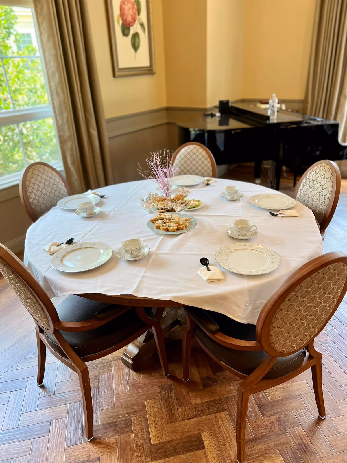 A round dining table set with a white tablecloth, six place settings with plates, teacups, spoons on napkins, and a centerpiece with pastries and decorative pink twigs. The room has wooden parquet flooring, beige walls, a window with curtains, a framed floral picture, and a black grand piano in the background.