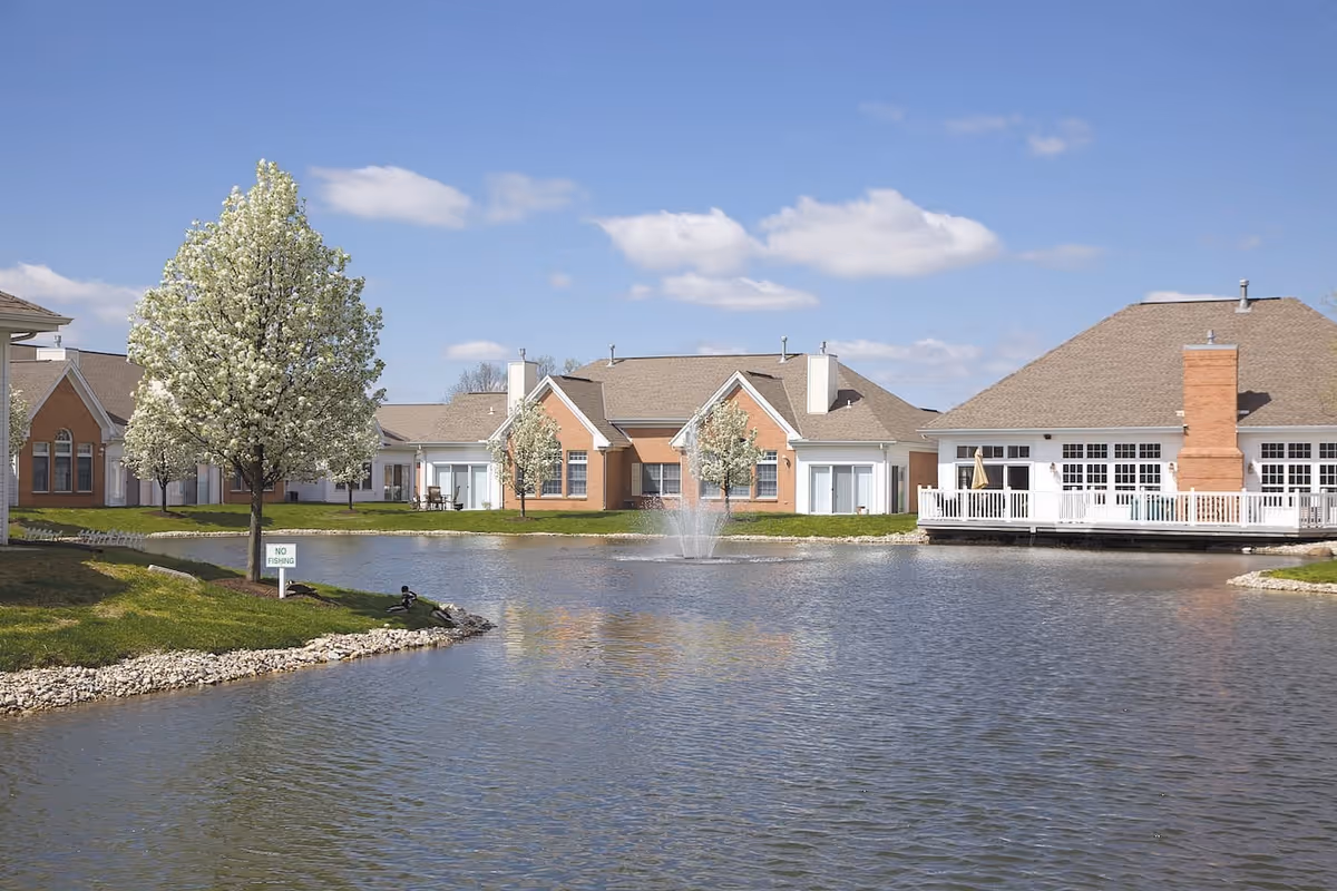 View of a senior living facility with a pond in the foreground, a water fountain in the pond, blooming trees along the grassy banks, and single-story buildings with brick and white siding under a blue sky with scattered clouds.