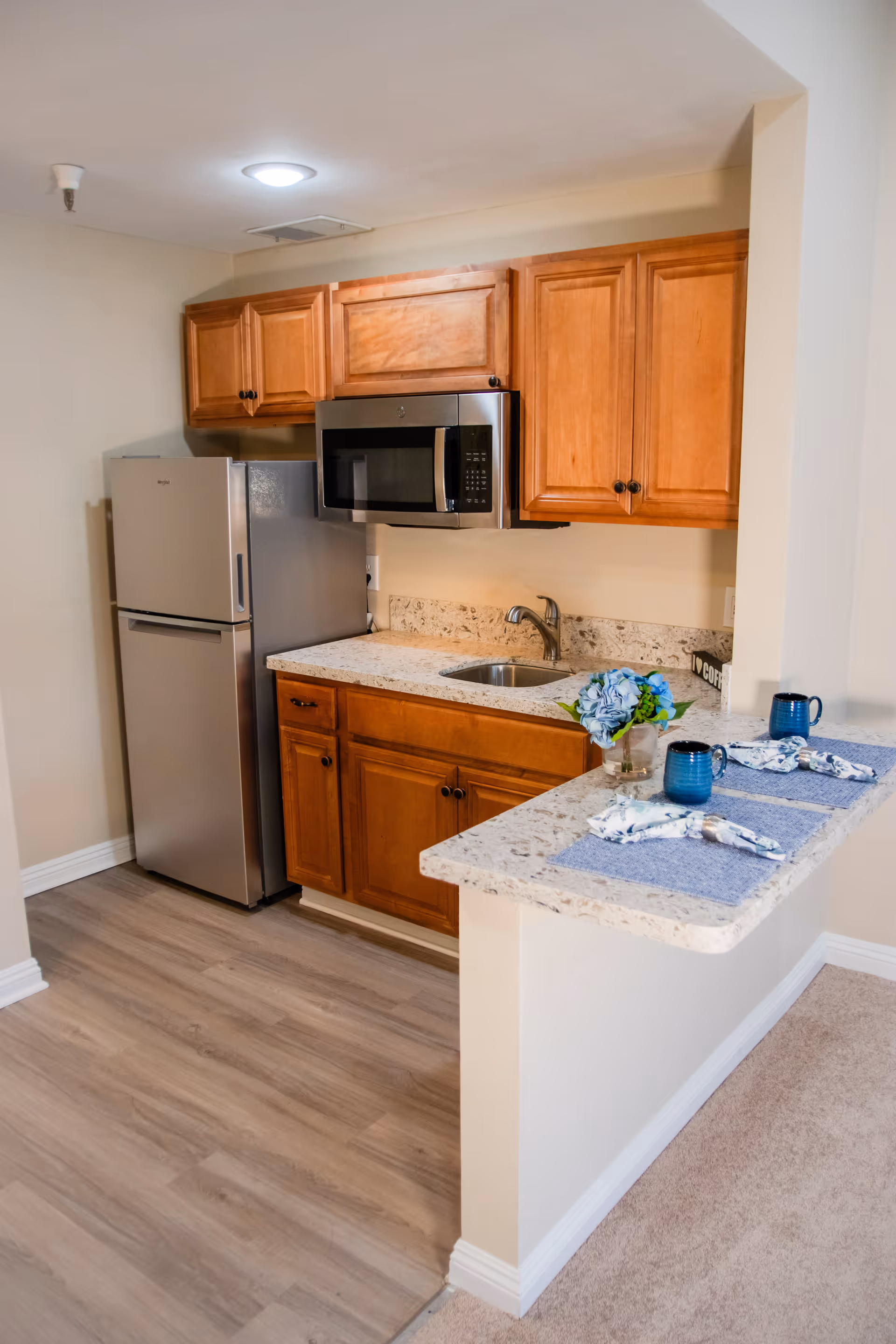 A small kitchen area with wooden cabinets, a stainless steel refrigerator, a microwave, a sink, and a granite countertop with two place settings including blue mugs and napkins. There is a small flower arrangement on the counter.