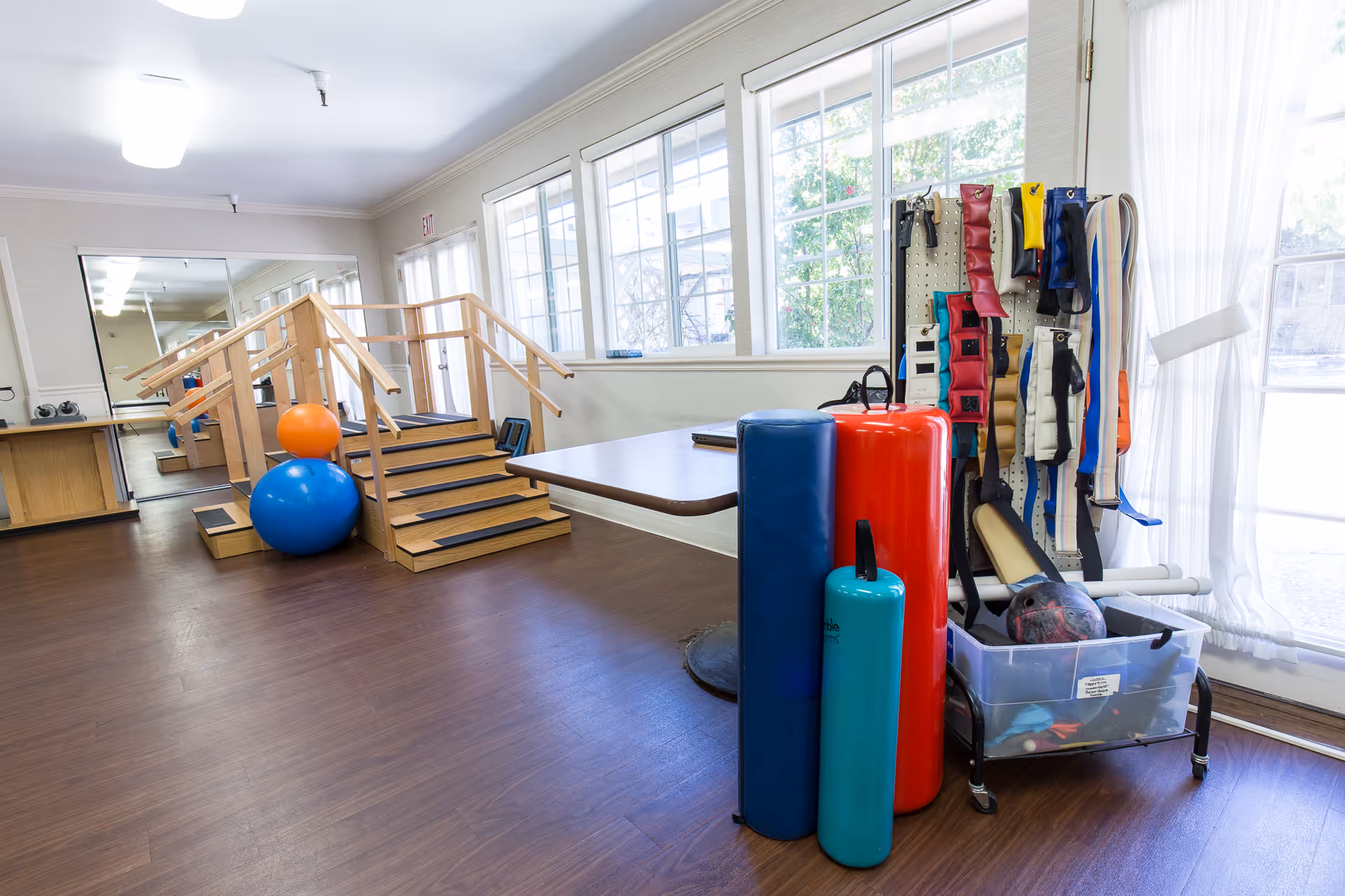 A physical therapy or rehabilitation room with wooden stairs for practice, large blue and orange exercise balls, various foam rollers in blue, red, and teal, and a rack holding multiple weighted belts and exercise equipment. The room has large windows letting in natural light and a wooden floor.