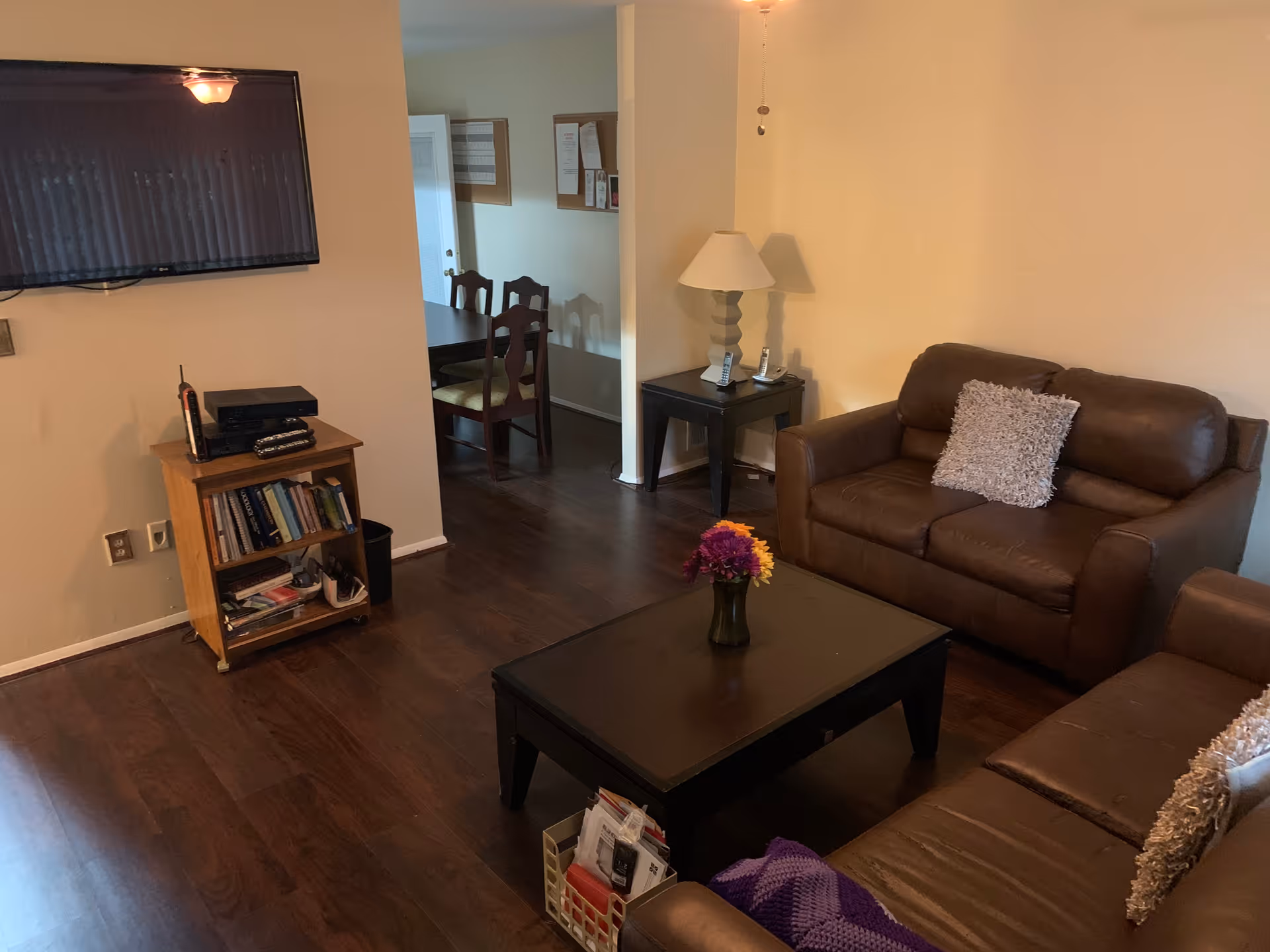 A cozy living room with two brown leather sofas, each with a textured throw pillow. A dark wooden coffee table with a small vase of colorful flowers sits in the center. To the left, a small wooden bookshelf holds books and a few items, with a flat-screen TV mounted on the wall above it. In the background, a dining area with a dark wooden table and chairs is visible. A side table with a lamp and two cordless phones is positioned between the sofas and the dining area. The room has wooden flooring and beige walls.