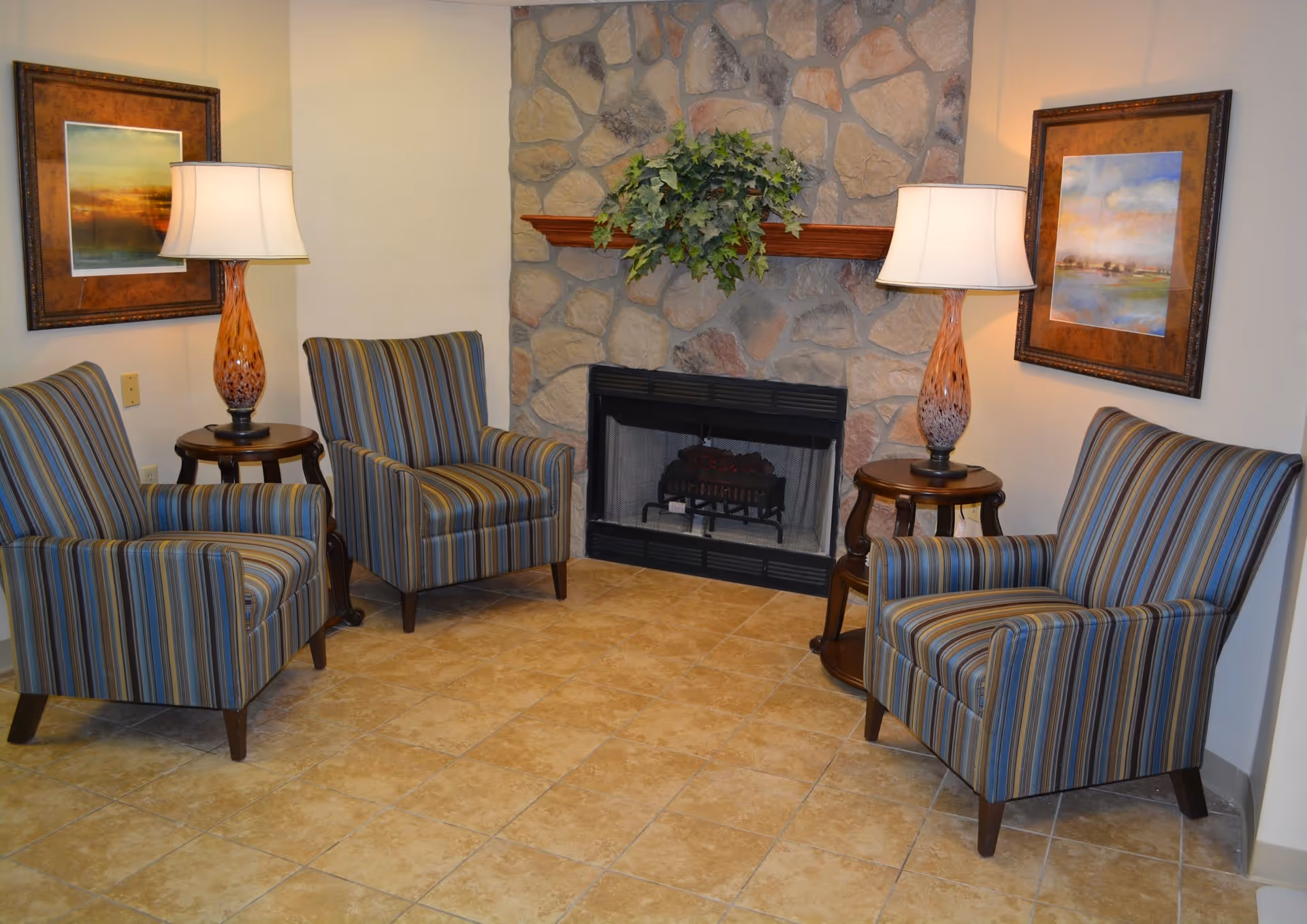 A cozy seating area with three striped armchairs arranged around a stone fireplace. Two wooden side tables each hold a decorative lamp with a white shade. Above the fireplace is a wooden mantel with a green leafy plant. Two framed landscape paintings hang on the walls on either side of the fireplace. The floor is tiled with light brown tiles.