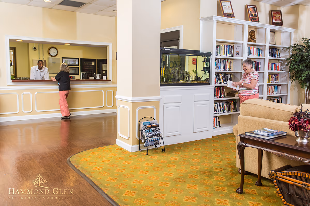 A bright and welcoming interior of a retirement community featuring a reception desk where a staff member is assisting a resident. To the right, another resident is browsing books on a white bookshelf next to a large fish tank. The room has a patterned carpet, wooden flooring, and comfortable seating with a coffee table and decorative flowers.