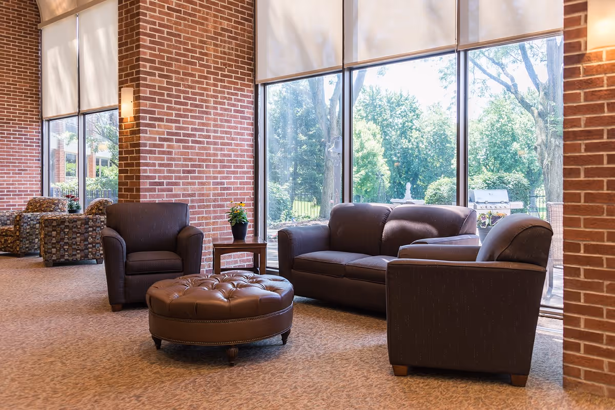 A cozy senior living facility lounge area with large floor-to-ceiling windows letting in natural light. The room features a brown leather sofa, two matching armchairs, a round tufted leather ottoman, and a small wooden side table with a potted plant. The walls are made of red brick, and outside the windows, green trees and a patio area with a grill are visible.
