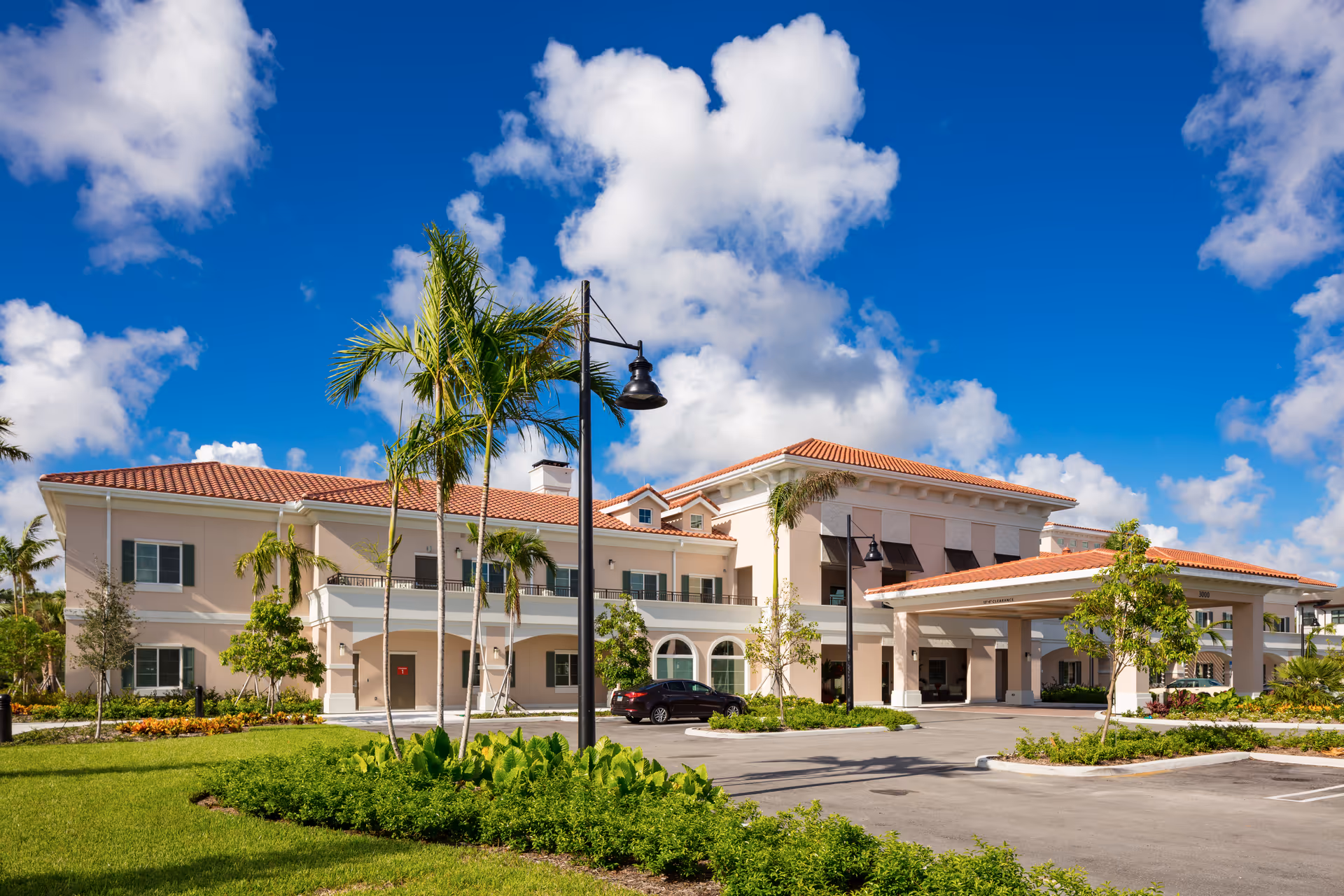 Exterior view of HarborChase of Palm Beach Gardens, a two-story building with a red-tiled roof, beige walls, and multiple windows. The entrance features a covered drop-off area with columns. The surrounding area includes palm trees, green lawns, and a clear blue sky with scattered clouds.