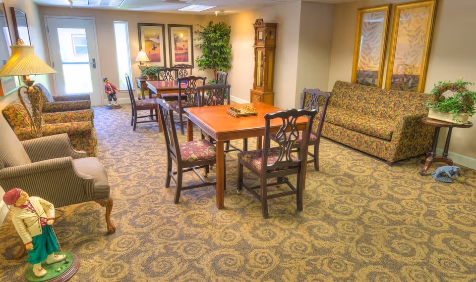 A cozy common area with patterned carpet, two wooden tables each surrounded by four chairs with floral upholstery, a floral-patterned sofa, two armchairs, a tall grandfather clock, framed artwork on the walls, potted plants, and decorative figurines including a golfer statue near the entrance door.