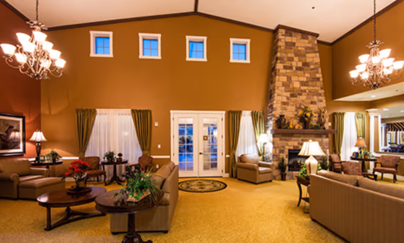 A spacious and warmly lit living room area with beige sofas and armchairs arranged around wooden coffee tables. The room features a tall stone fireplace, multiple lamps, chandeliers, and windows with green curtains. The walls are painted in warm tones, and there is a double glass door in the center with small windows above it.