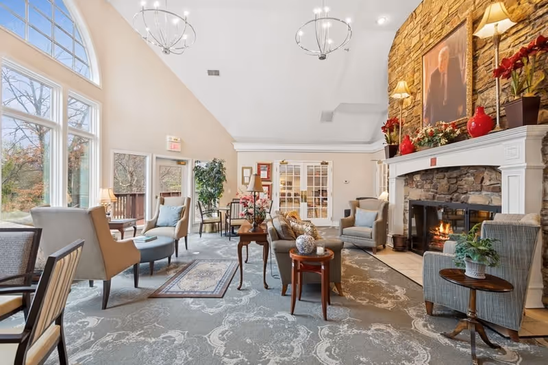 A spacious and well-lit living room area with large windows letting in natural light. The room features comfortable armchairs and sofas arranged around a stone fireplace with a fire burning. The fireplace mantel is decorated with red vases, flowers, and lamps. There are side tables with plants and decorative items, a patterned carpet, and two modern chandeliers hanging from the ceiling. French doors and a door leading outside are visible in the background.