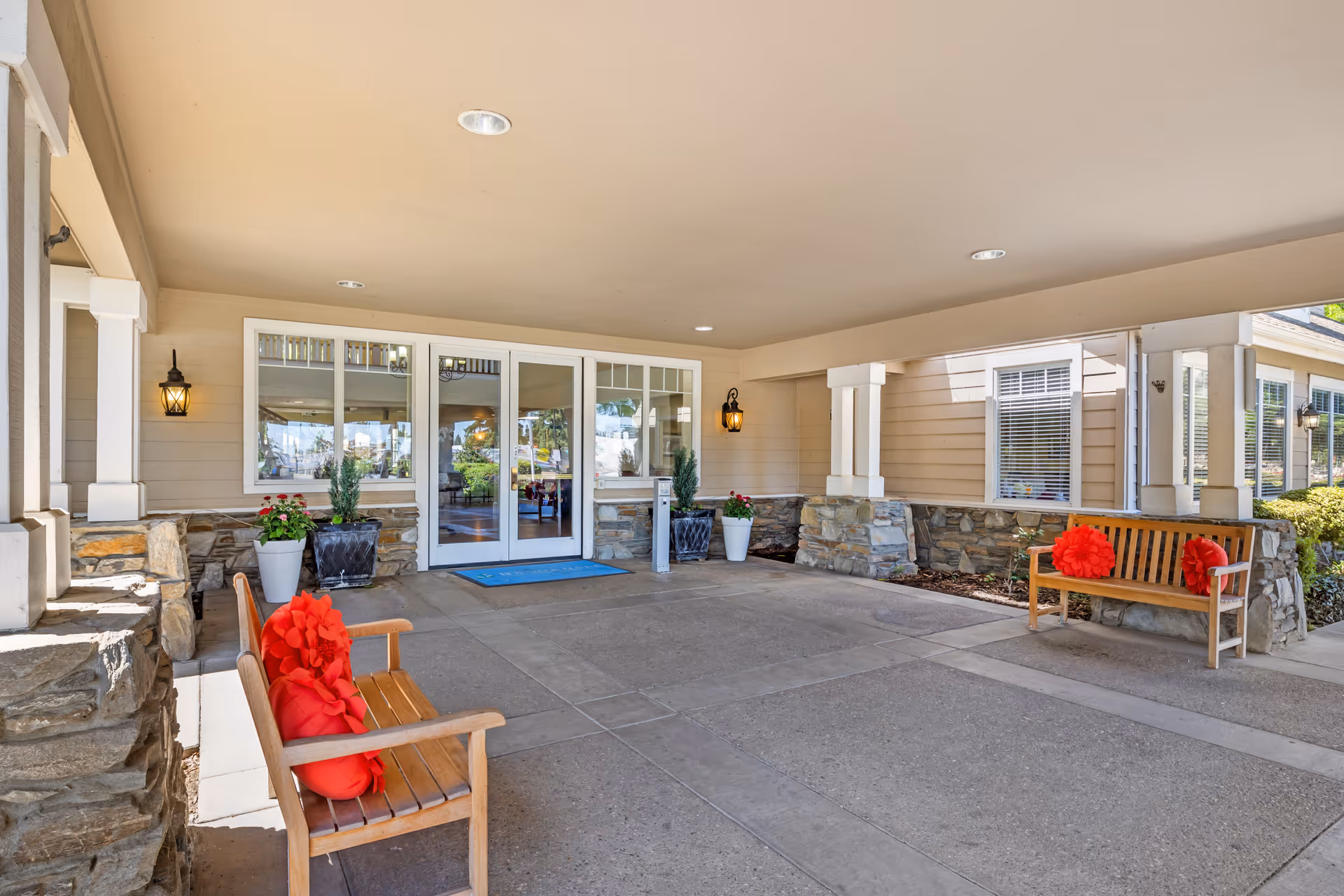 Covered entrance area of a senior living facility with two wooden benches, each adorned with red flower-shaped cushions. The entrance has double glass doors with windows on either side, stone accents on the lower walls, potted plants, and wall-mounted lantern-style lights.