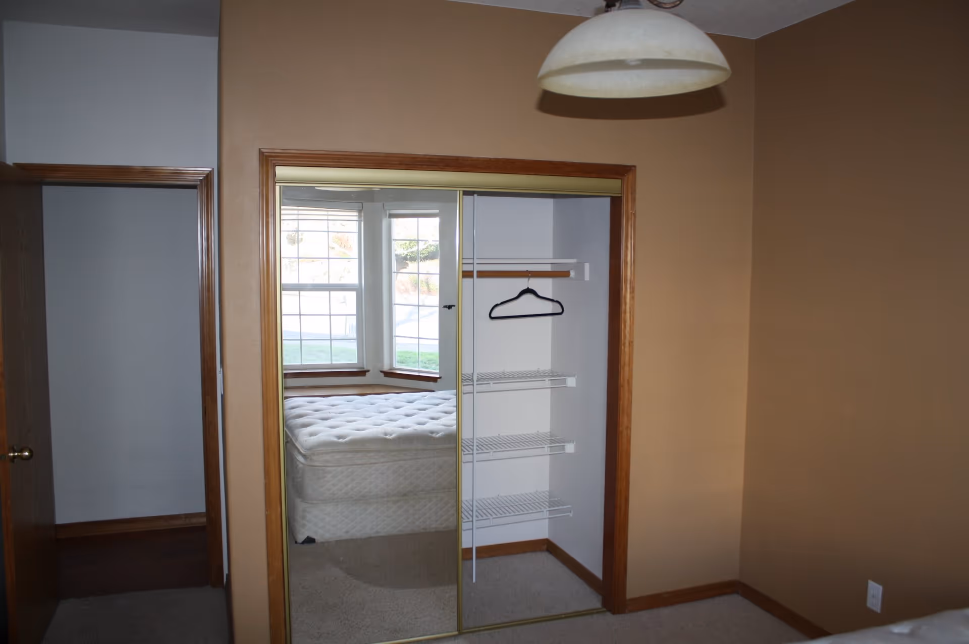 Interior view of a bedroom with beige walls and carpeted floor. There is a closet with sliding mirrored doors, one side open showing wire shelves and a single black hanger. Through the mirror, a bed with a white mattress and a bay window with white blinds are visible. A ceiling light fixture hangs from above.