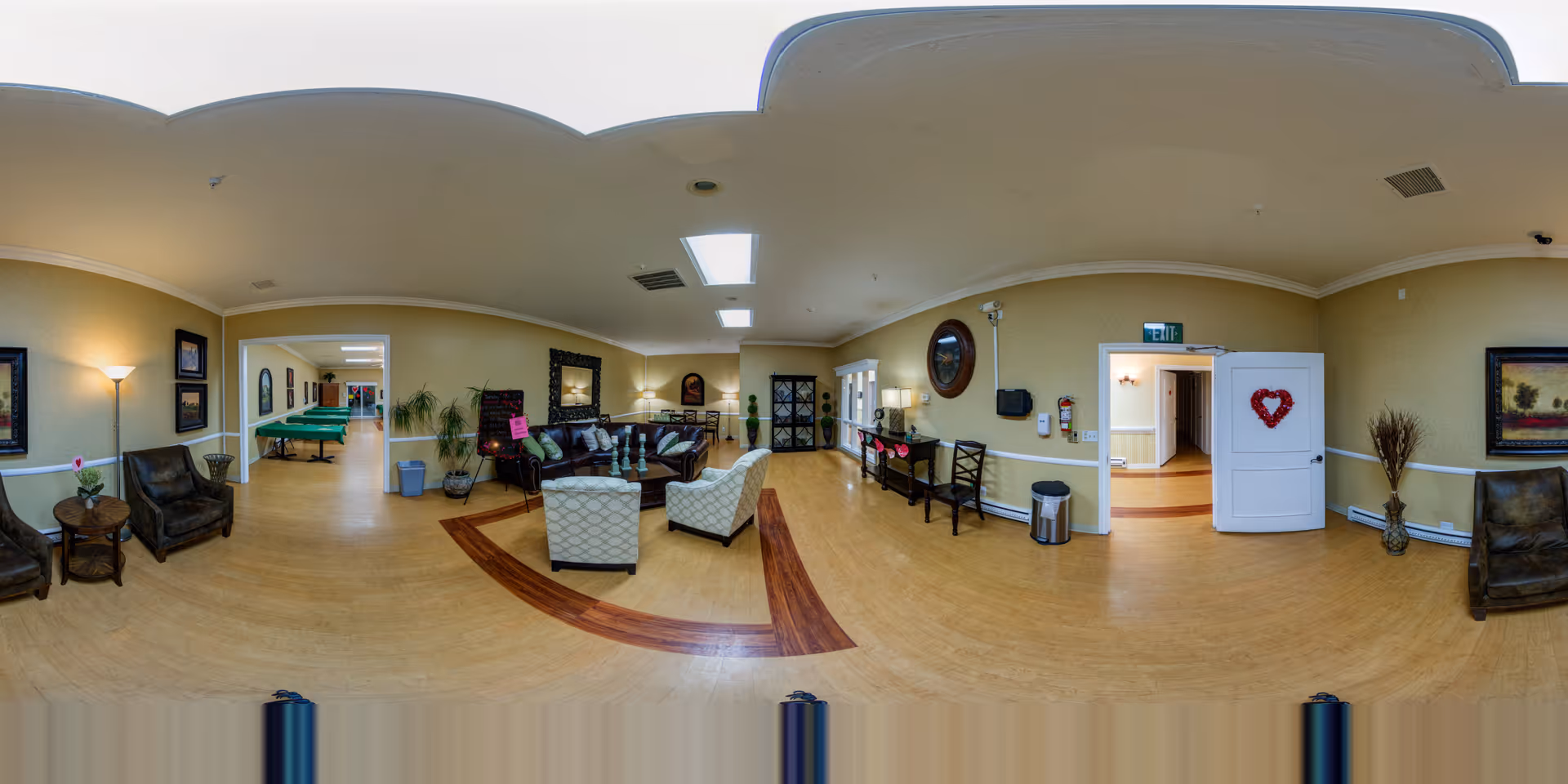 A spacious senior living facility common area with comfortable seating including armchairs and a sofa arranged around a coffee table. The room has light yellow walls, wooden flooring with a decorative inlay, and ceiling lights including skylights. There are framed pictures on the walls, a floor lamp, plants, and a doorway with a white door decorated with a red heart-shaped wreath. In the background, a hallway and a pool table are visible.