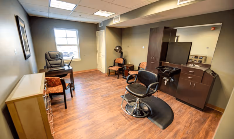 A well-lit room with wood flooring featuring a black salon chair on a mat in front of a dark wood cabinet with a large mirror and a sink. There are two orange cushioned chairs and a small table against the wall, a desk with a black office chair near a window, and a framed picture on the wall.