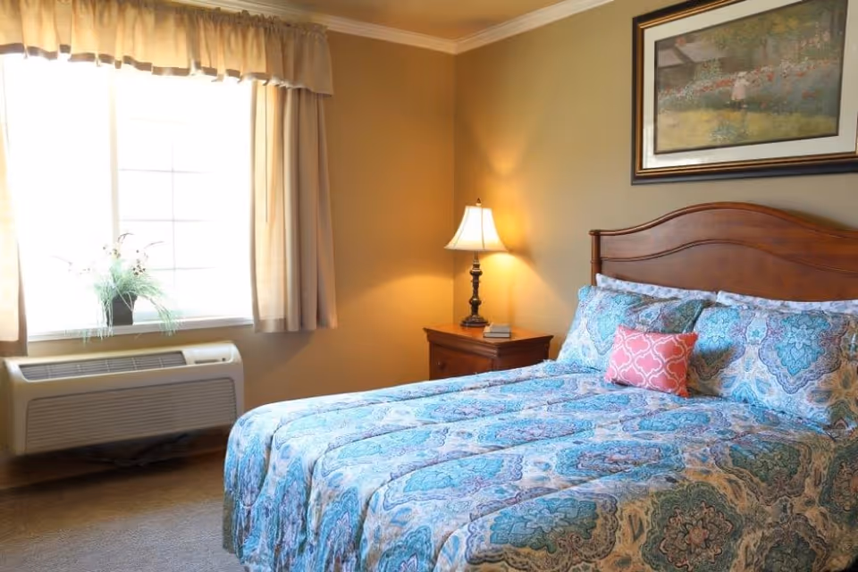 A cozy bedroom with a large bed covered in a blue and white patterned comforter and pillows, including a small pink decorative pillow. There is a wooden nightstand with a lamp and a small book next to the bed. A window with beige curtains allows natural light into the room, and a framed painting hangs on the wall above the bed.