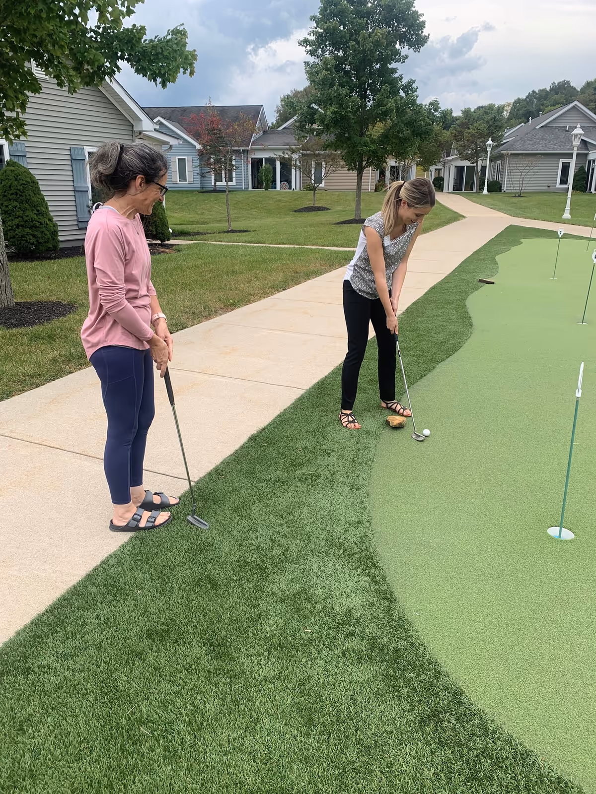Two women playing mini golf on a putting green outside in a residential community area with houses and trees in the background. One woman is preparing to putt while the other watches.