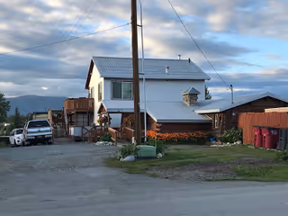 A two-story white building with a metal roof, wooden stairs on the side, and a small porch area. There are several red trash bins lined up near a wooden shed on the right side. A white pickup truck is parked on the left side of the building. The sky is partly cloudy with mountains visible in the background.