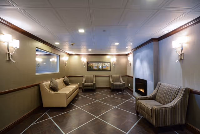 A cozy interior lounge area with a beige sofa, three armchairs, a fireplace on the right wall, and a framed landscape painting on the far wall. The room has soft lighting from wall sconces and a ceiling with recessed lights. The floor is dark wood with a geometric pattern.