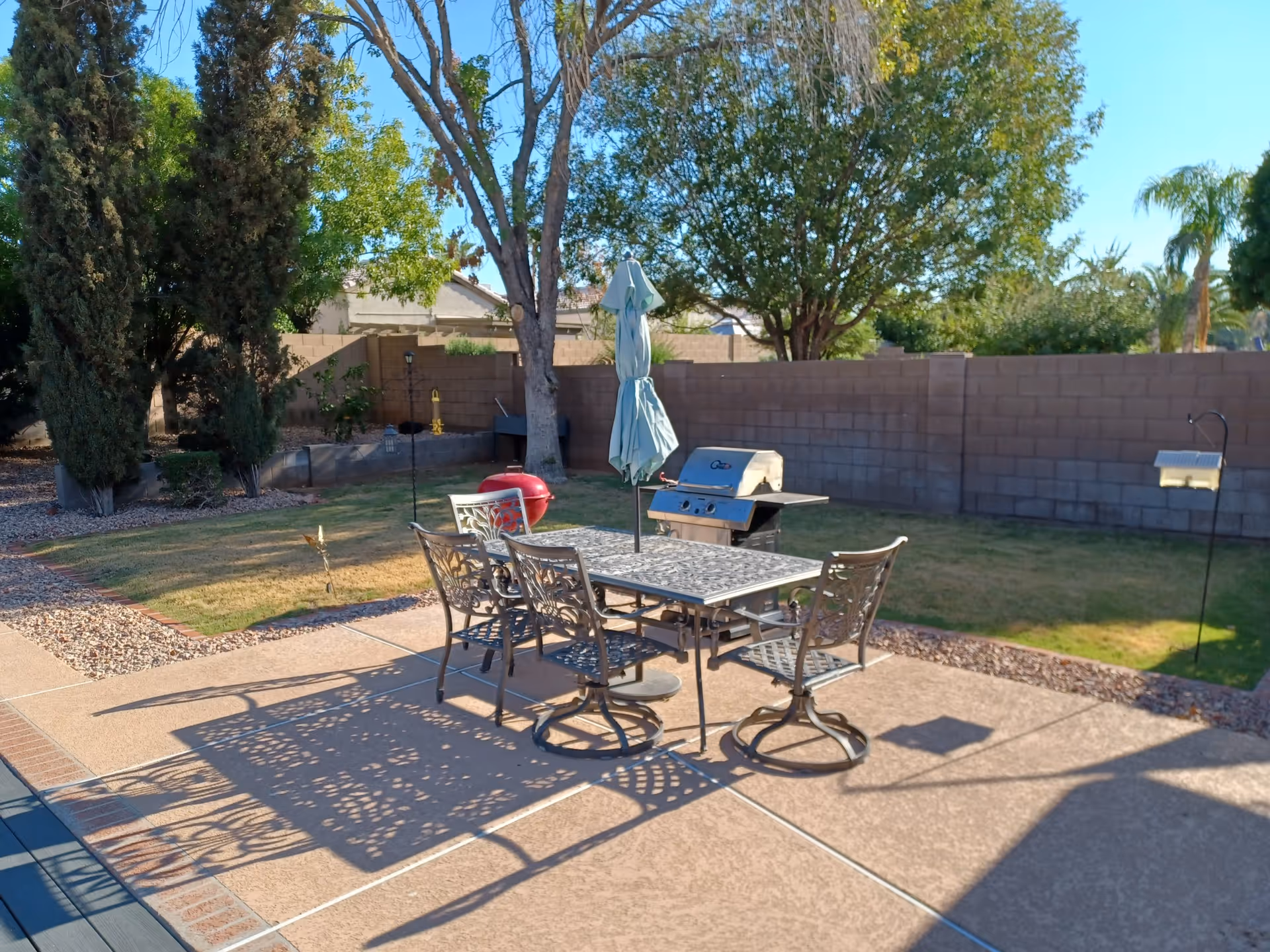 Outdoor patio area with a metal table and four chairs, a closed umbrella in the center of the table, a barbecue grill, and a red kettle grill. The patio is surrounded by grass, trees, and a brick wall fence under a clear blue sky.