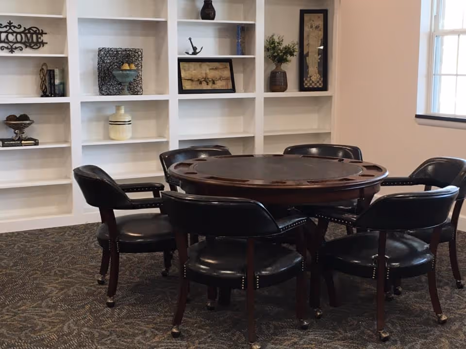 A round wooden table with six black leather chairs arranged around it in a room with white built-in shelves displaying decorative items and a window on the right side.