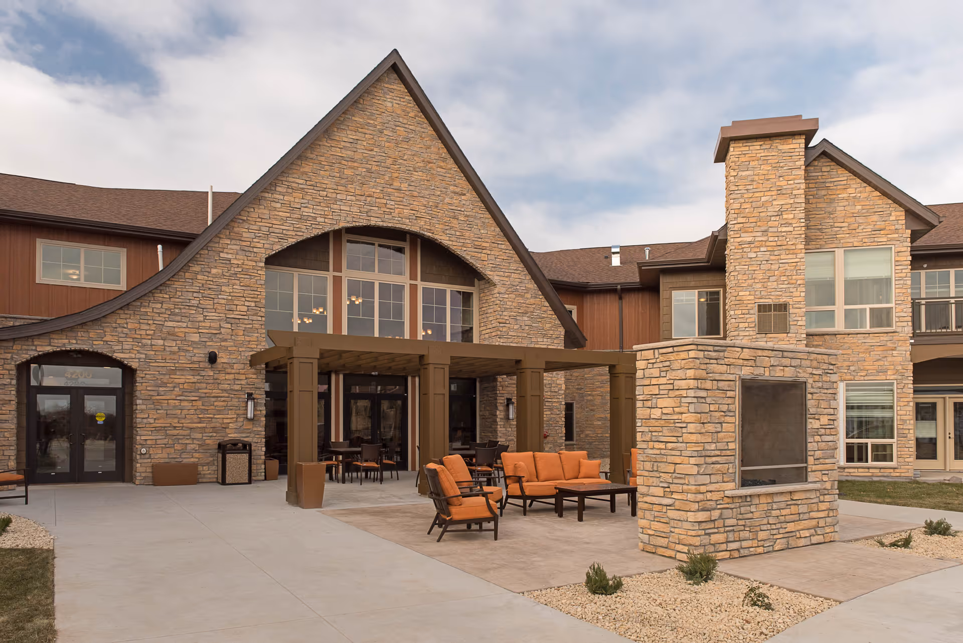 Outdoor patio area of a senior living facility with stone exterior walls, a large stone fireplace, and orange cushioned seating including chairs and a sofa. The building has large windows and a covered seating area under a wooden pergola.
