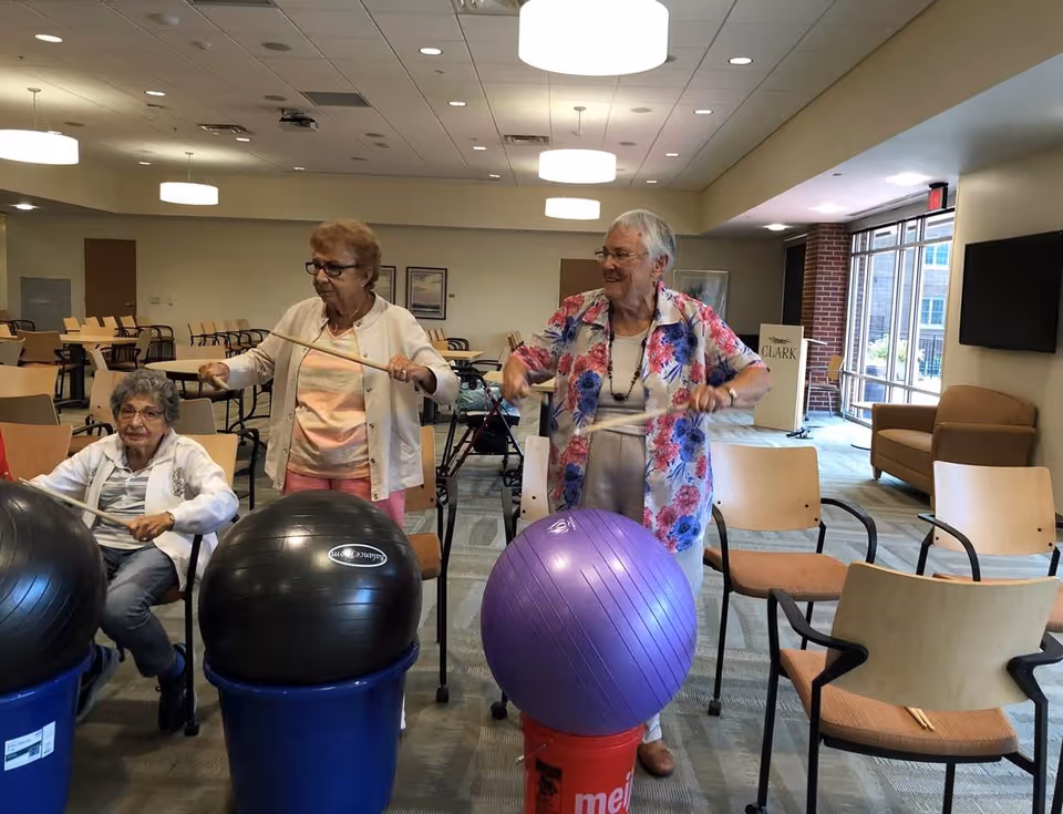 Three elderly women participating in a seated exercise activity using drumsticks to hit large exercise balls placed on buckets in a spacious room with chairs and large windows.