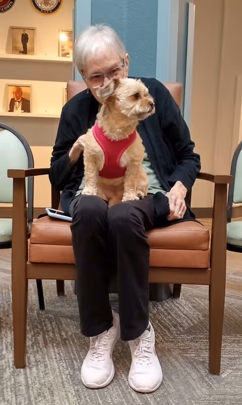 An elderly person with short white hair and glasses sits on a brown leather chair indoors, holding a small dog wearing a red harness on their lap. The background shows a light blue wall and shelves with framed photos.