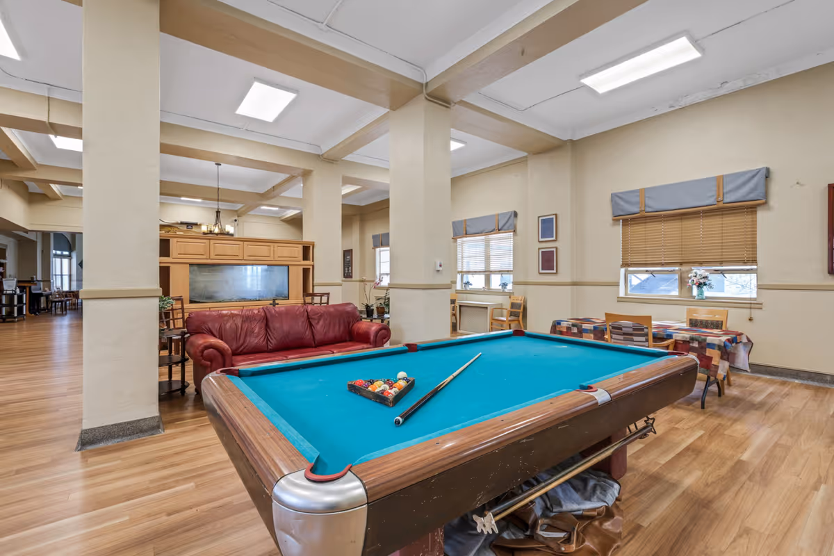 Interior view of a senior living facility common area featuring a pool table with a cue stick and racked balls, a red leather couch behind the pool table, wooden flooring, beige walls with windows covered by blinds and valances, and tables with colorful tablecloths and chairs near the windows.