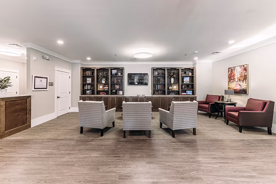 A spacious senior living facility common area with a row of three patterned armchairs facing built-in wooden bookshelves and a TV mounted on the wall displaying 'South High Senior Living'. To the right, there are two maroon armchairs with a small round table and lamp between them, and a framed picture of a tree-lined path on the wall. The floor is a combination of wood and carpet, and the walls are painted light beige with white trim.