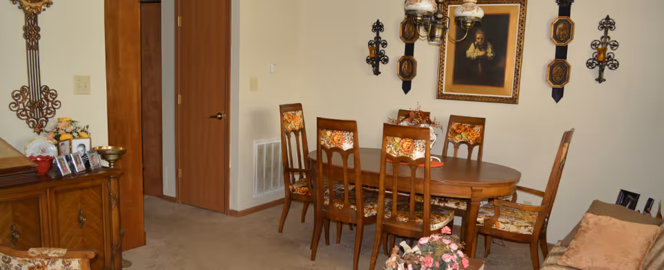 A dining room with a wooden oval table surrounded by six wooden chairs with floral upholstery. The walls are decorated with framed artwork and ornate wall sconces. There is a wooden sideboard with framed photos and decorative items on the left side. The room has beige carpet and light-colored walls.